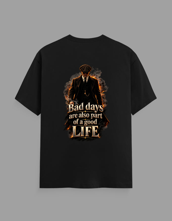 A black short-sleeve t-shirt with a graphic print on the front, featuring a dark figure in a suit surrounded by flames and the text 'Bad days are also part of a good LIFE'. The t-shirt is displayed flat against a solid gray background.