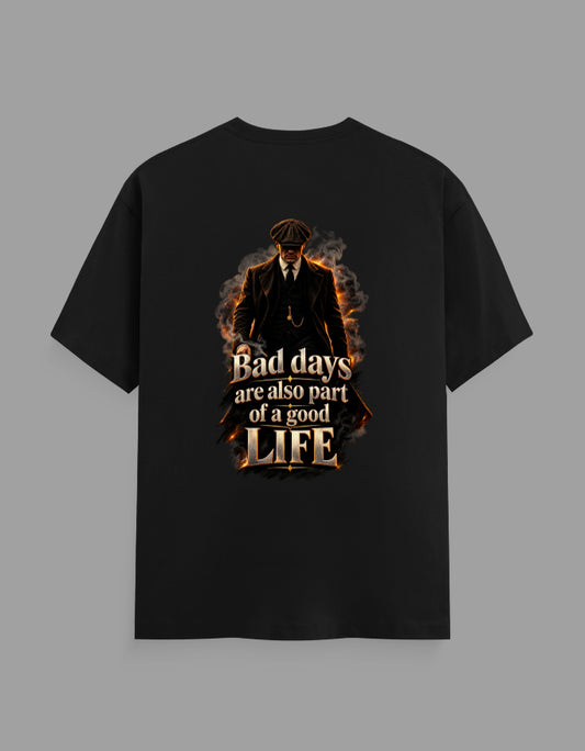 A black short-sleeve t-shirt with a graphic print on the front, featuring a dark figure in a suit surrounded by flames and the text 'Bad days are also part of a good LIFE'. The t-shirt is displayed flat against a solid gray background.